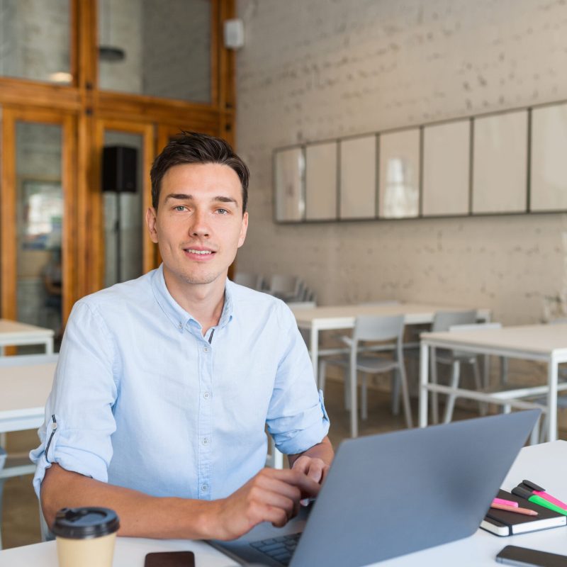 young handsome smiling man sitting in open space office working on laptop, happy, positive, successful modern youth, online education