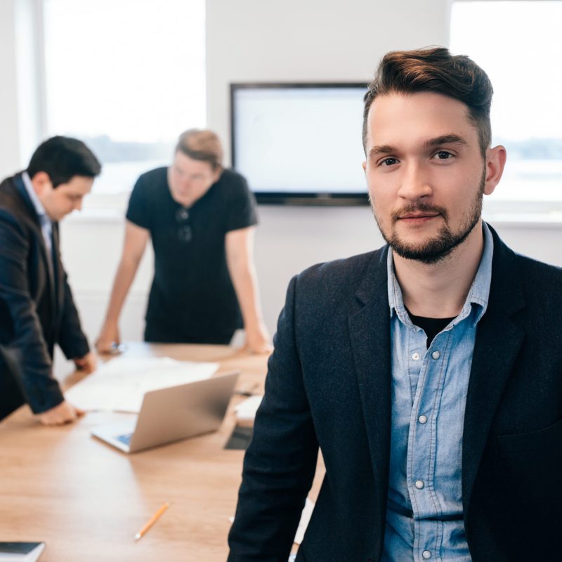 Two colleagues are working  at  table in the office. They wear black clothes. There is young guy looking to the camera on front.