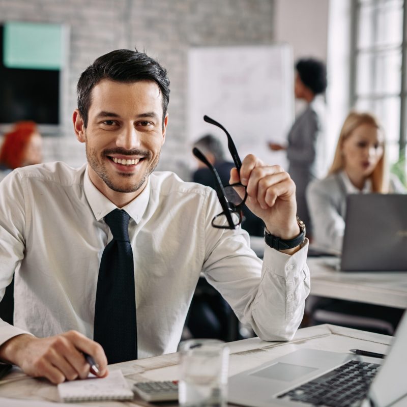 Young happy businessman working in the office and looking at camera. There are people in the background.